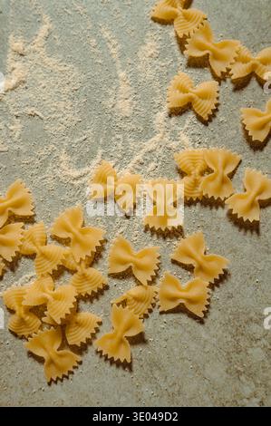 Un délicieux arrangement de pâtes farfalle italiennes crues dispersées sur une surface rustique saupoudrée de farine. La forme de noeud papillon des pâtes ajoute charme et Banque D'Images