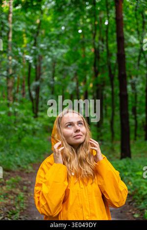 Une femme portant un imperméable jaune vif se tient dans une forêt luxuriante et verte. Elle regarde vers le haut, peut-être à l'écoute des oiseaux, embrassant l'ambiance tranquille i. Banque D'Images