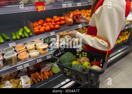Femme choisissant des tomates dans l'allée de l'épicerie. Un acheteur sélectionne des tomates fraîches dans une section de produits de supermarché tout en transportant un panier rempli de Banque D'Images