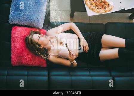 Une femme est allongée sur un canapé, tenant un joint de marijuana avec désinvolture. Elle semble détendue et confortable, entourée d'oreillers colorés, suggérant un laid-b Banque D'Images