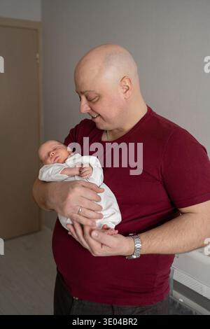 Un père tient tendrement son nouveau-né dans un environnement familial confortable. Le nourrisson dort paisiblement dans une combinaison blanche, mettant en valeur la chaleur et l'amour Banque D'Images