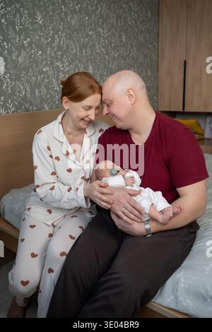 Une scène réconfortante d'une famille aimante à la maison avec leur nouveau-né. Les parents, assis ensemble sur un lit, partagent un moment tendre en berçant le leur Banque D'Images
