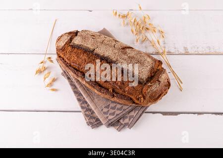 Pain de grain entier fraîchement cuit reposant sur une serviette à motifs. De délicates tiges de grains accompagnent l'arrangement sur une table en bois blanc rustique Banque D'Images
