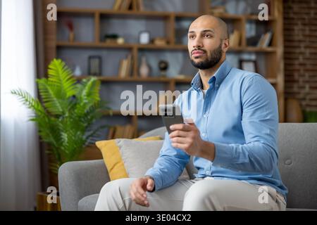 Homme réfléchi assis sur un canapé, tenant un téléphone portable et regardant loin, réfléchissant à de nouvelles idées ou des solutions de travail à distance dans un salon confortable Banque D'Images