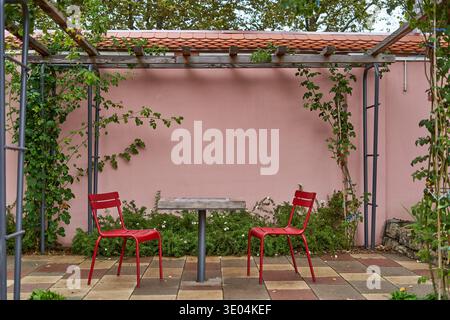 Une scène de patio extérieur sereine avec deux chaises en métal rouge vif se faisant face sur une petite table en bois, contre un mur rose pastel.... Banque D'Images