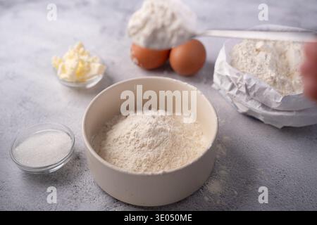 gros plan de préparations de cuisson sur un comptoir texturé gris : un bol en céramique empilé de farine, œufs, beurre, sucre et un sac en papier froissé, journée douce Banque D'Images