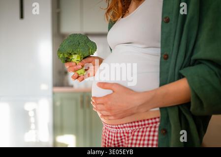 Attendre une femme embrassant sa grossesse, tenant un fleuron de brocoli frais, symbolisant des habitudes alimentaires saines et incorporant un régime nutritif pour le bien-être prénatal et le développement du bébé Banque D'Images