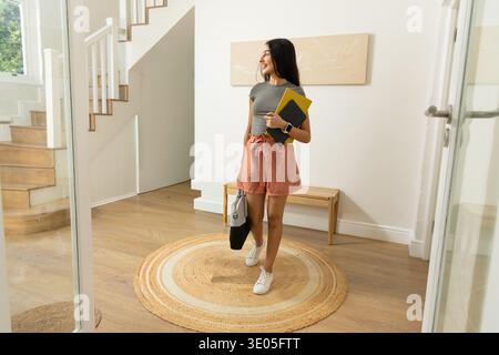 Femme indienne au début des années 20 marchant dans le hall portant des baskets blanches tenant deux cahiers et un fourre-tout. Entrée, lumière du jour, bois dur, escalier, banc, mur Banque D'Images