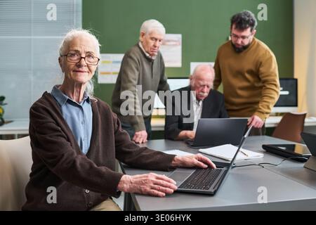 Portrait d'une femme caucasienne senior utilisant un ordinateur portable pendant un cours d'alphabétisation informatique avec des hommes caucasiens seniors et un homme caucasien d'âge moyen aidant dans un cadre de classe Banque D'Images