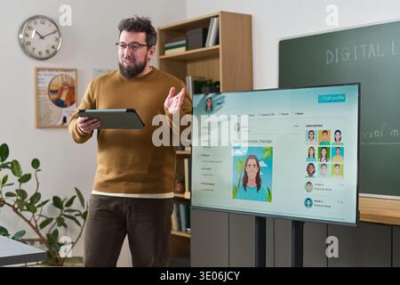 Homme d'âge moyen caucasien debout dans la salle de classe tenant une tablette numérique présentant le contenu du cours d'alphabétisation informatique sur un grand moniteur avec des profils d'utilisateur illustrés visibles Banque D'Images