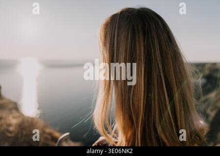 Femme cheveux coucher de soleil voyageur se tient sur la côte regardant le reflet doré sur l'océan au cours d'une soirée tranquille Banque D'Images