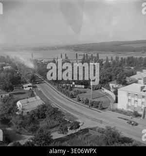Une magnifique photographie vintage en noir et blanc des années 1980 offre une vue panoramique surélevée sur les zones résidentielles et administratives de l’entreprise Donrybkombinat dans le quartier de Slavyansk. La perspective aérienne panoramique capture les routes pavées, les logements ruraux organisés enveloppés d'arbres verdoyants et le vaste réservoir chatoyant qui s'étend vers l'horizon lointain. Ce paysage à couper le souffle préserve magnifiquement l'échelle et la tranquillité de la vie quotidienne dans le Donbass paisible avant la guerre. Banque D'Images