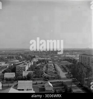 Une vaste photographie vintage en noir et blanc des années 1980 présente une vue imprenable sur le quartier résidentiel et culturel central de l'entreprise Donrybkombinat dans le quartier de Slavyansk. La perspective aérienne panoramique capture l'éminente Maison de la culture, les immeubles d'habitation environnants et les arbres verdoyants avec pour toile de fond un vaste réservoir tranquille. Ce paysage authentique préserve magnifiquement l'infrastructure communautaire et la vie quotidienne dans le Donbass paisible avant la guerre. Banque D'Images