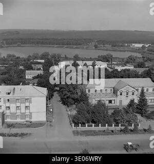 Une vaste photographie vintage en noir et blanc des années 1980 présente une vue imprenable sur le quartier résidentiel et culturel central de l'entreprise Donrybkombinat dans le quartier de Slavyansk. La perspective aérienne panoramique capture l'éminente Maison de la culture, les immeubles d'habitation environnants et les arbres verdoyants avec pour toile de fond un vaste réservoir tranquille. Ce paysage authentique préserve magnifiquement l'infrastructure communautaire et la vie quotidienne dans le Donbass paisible avant la guerre. Banque D'Images