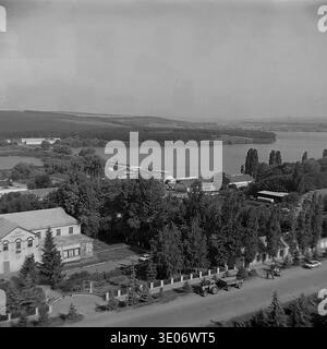 Une vaste photographie vintage en noir et blanc des années 1980 présente une vue imprenable sur le quartier résidentiel et culturel central de l'entreprise Donrybkombinat dans le quartier de Slavyansk. La perspective aérienne panoramique capture l'éminente Maison de la culture, les immeubles d'habitation environnants et les arbres verdoyants avec pour toile de fond un vaste réservoir tranquille. Ce paysage authentique préserve magnifiquement l'infrastructure communautaire et la vie quotidienne dans le Donbass paisible avant la guerre. Banque D'Images