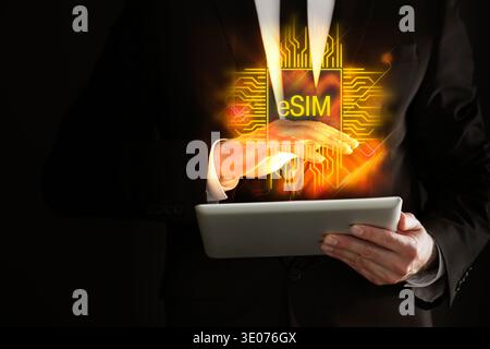 Homme utilisant une tablette sur fond noir, gros plan. Carte SIM électronique virtuelle (eSIM) au-dessus de l'appareil Banque D'Images