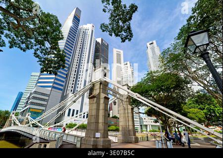 Les gens traversent un pont dans la zone financière de Singapour. De grands bâtiments entourent la zone par temps clair avec des arbres verts à proximité. Banque D'Images