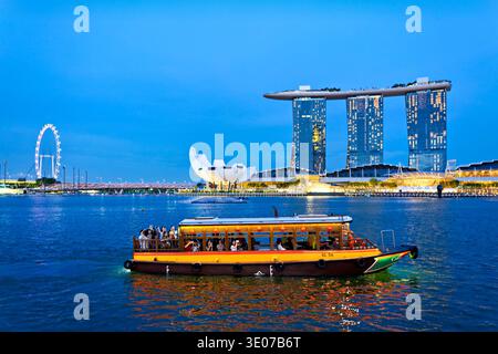 Un bateau voyage sur l'eau près de Merlion et Marina Bay Sands tandis que les gens profitent de la vue du coucher du soleil sur la ligne d'horizon de Singapour. Banque D'Images