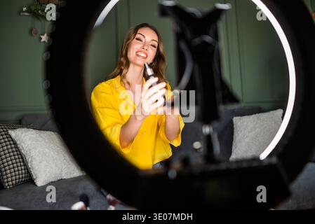 Une femme est assise sur un canapé enregistrant un tutoriel beauté avec smartphone, éclairé par une lumière annulaire. Banque D'Images