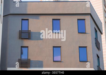 L'extérieur d'un immeuble résidentiel contemporain présente une façade grise texturée avec de multiples fenêtres teintées de bleu et des balcons minimalistes baignés Banque D'Images