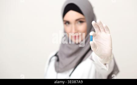 Femme en manteau blanc tient la capsule bleue tout en portant Hijab et gants dans le cadre médical Banque D'Images