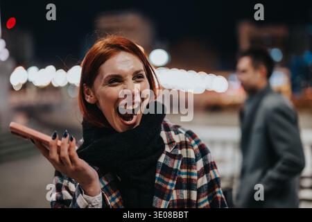 Femme riante avec téléphone rose à l'extérieur la nuit dans un manteau à carreaux et une écharpe Banque D'Images
