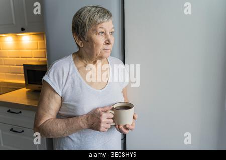 Femme senior regardant par la fenêtre lumineuse, tenant une tasse à café, pensant à la vie ou à l'avenir dans un moment calme Banque D'Images