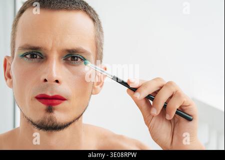 homme gay caucasien adulte avec maquillage et cosmétiques sur le visage. Homme transgenre avec rouge à lèvres rouge appliqué ombre à paupières scintillant sur ses yeux avec un pinceau sur un fond blanc Banque D'Images