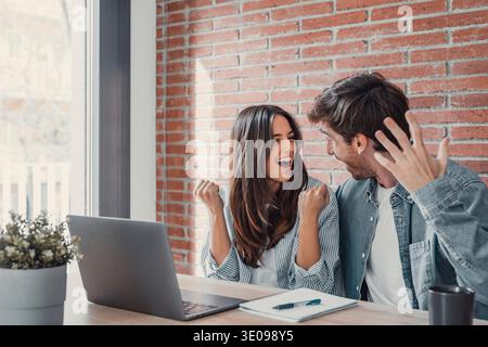 Un couple millennial ravi assis au bureau regarder l'écran d'ordinateur portable se sentir euphorique avec la victoire en ligne, heureux jeune mari et femme excité lire de bonnes nouvelles, obtenir Banque D'Images