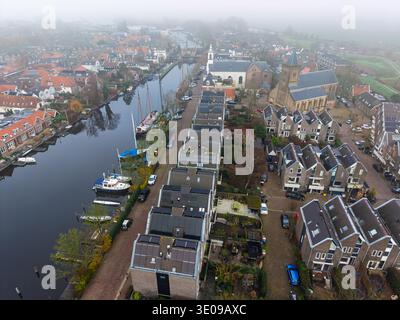 Vue aérienne d'une ville néerlandaise brumeuse avec un canal, une église historique et des maisons modernes avec panneaux solaires. Une scène matinale d'hiver sombre mettant en vedette des bateaux et Banque D'Images