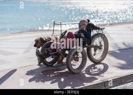 L'homme monte un tricycle allongé adaptatif avec son chien le long d'une promenade ensoleillée en bord de mer, mettant en évidence les loisirs en plein air accessibles et l'inclusion de la mobilité. Alicante, Espagne - 11 juin 2025 Banque D'Images