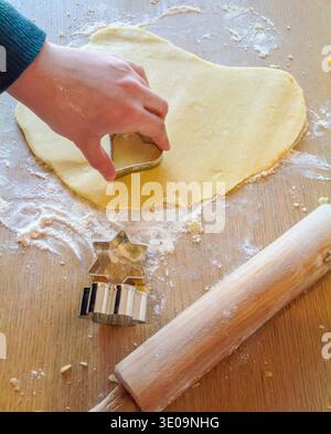 Enfant utilisant des emporte-pièces en forme de coeur sur la pâte, avec rouleau à pâtisserie et emporte-pièces en forme d'étoile sur la table en bois, faisant des biscuits Banque D'Images
