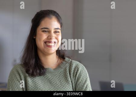 Femme souriante tout en étant assise dans le bureau, portant un pull en maille vert sauge et ordinateur portable visible Banque D'Images