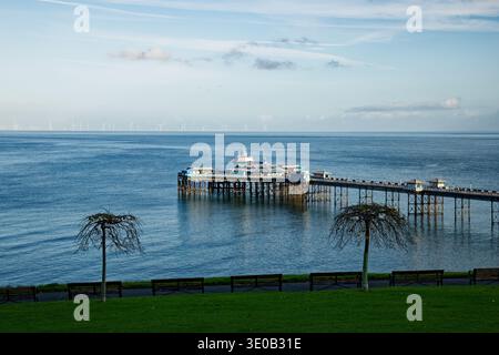 Une ligne de bancs de parc vides avec deux arbres en forme de champignons sans feuilles surplombe une mer bleue et un quai à Llandudno, au nord du pays de Galles. Banque D'Images