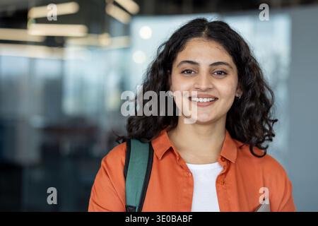 Jeune femme indienne aux cheveux bouclés portant un sac à dos et une tenue décontractée, souriant joyeusement tout en profitant de sa vie universitaire et en poursuivant une éducation réussie dans un cadre universitaire moderne Banque D'Images