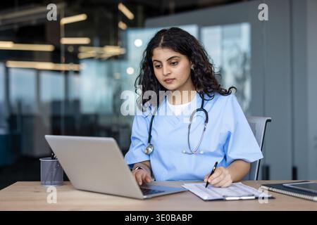 Jeune spécialiste médicale indienne portant des gommages bleus et un stéthoscope, se concentrant sur ses notes et son ordinateur portable tout en travaillant à un bureau dans un environnement hospitalier moderne Banque D'Images