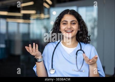Jeune professionnelle médicale indienne dans les gommages bleus et stéthoscope souriant, parlant et faisant des gestes avec les mains, symbolisant la communication, les soins et le professionnalisme en matière de soins de santé Banque D'Images