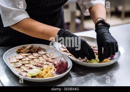 Gros plan des mains d'un chef qui dispose de tranches de viande, de jambon et de petits pains sur des assiettes blanches. L'apéritif est garni de concombres et de sauce. Prépar culinaire Banque D'Images