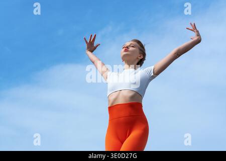 Plan bas angle d'une jeune femme sereine en leggings orange vif et un haut blanc. Elle se tient debout avec les bras tendus vers le ciel bleu, embrassant une sensation Banque D'Images