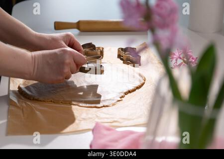Les mains façonnent la pâte à pain d'épices à l'aide d'emporte-pièces sur une table de cuisine. Rouleau à pâtisserie et fleurs sont à proximité. Cette activité fait partie de la préparation de Pâques et de ses traditions. Banque D'Images