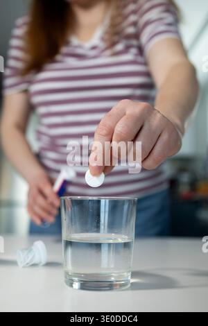 Une personne laisse tomber un comprimé effervescent blanc dans un verre d'eau claire sur une surface blanche propre. La personne porte une chemise rayée et tient une bouteille en arrière-plan, préparant une boisson dissoute. Banque D'Images