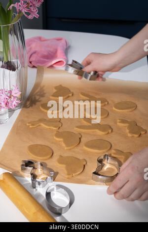 Les mains découpent la pâte à pain d'épices crue en formes de lapin de Pâques, d'œuf et de poussin sur du papier sulfurisé. Un vase de jacinthes roses se trouve à proximité alors que les préparatifs pour la célébration des fêtes se poursuivent dans la cuisine. Banque D'Images