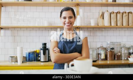 Café, comptoir et portrait de femme avec les bras croisés, boisson caféine ou cappuccino. Serveuse, barista et personne heureuse avec tasse dans Banque D'Images