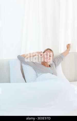 Femme allongée et étirée sur le lit dans la chambre portant un haut de pyjama gris clair avec des oreillers blancs. Minimaliste, serein, matin, beige, lin, coussins Banque D'Images
