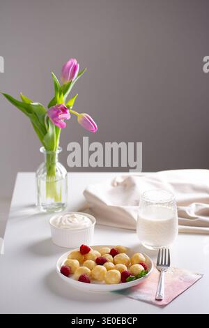 Boulettes paresseuses ukrainiennes à la crème sure et aux framboises fraîches servies sur une table blanche décorée de tulipes roses dans un intérieur lumineux. Banque D'Images