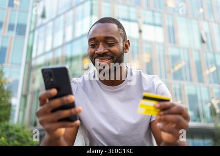 Homme africain magasinant en ligne tenant smartphone payant avec carte de crédit d'or. Guy sur la rue urbaine dans la ville achetant sur Internet entrer les détails de la carte de crédit. Service de livraison de commerce électronique d'achat en ligne Banque D'Images