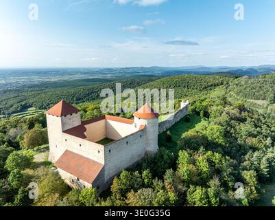 Vue aérienne d'une forteresse médiévale sur une colline avec des remparts en pierre blanche et des tours aux toits rouges, dominant une forêt verte dense, près d'Amberieu, Ain, France Banque D'Images