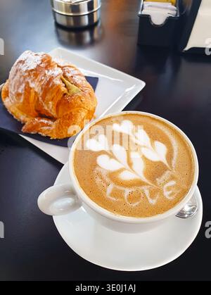 Tasse de cappuccino avec latte art mettant en vedette le mot « amour » et un motif de feuilles décoratif, servi avec un croissant frais dans un cadre de café moderne. Romantique Banque D'Images