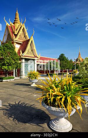 Les oiseaux volent au-dessus du Palais Royal à Phnom Penh. Des arbres verts entourent le palais. La lumière du soleil brille sur les toits dorés et les fleurs. Banque D'Images