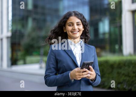 Une jeune femme professionnelle aux cheveux bouclés foncés sourit en toute confiance tout en tenant un smartphone à l'extérieur des immeubles de bureaux modernes, symbolisant la réussite de carrière urbaine et le réseautage numérique Banque D'Images